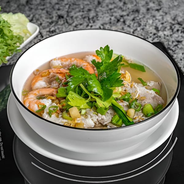 A white bowl of Vietnamese soup with shrimp, meat, green onions, and cilantro on a dark surface.